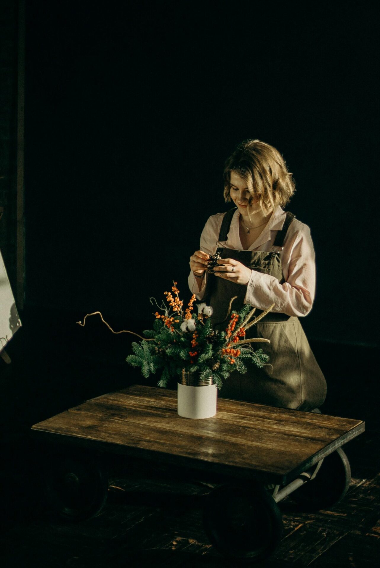 A woman arranges a floral bouquet on a wooden table in a dimly lit room, wearing an apron.